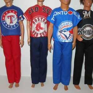 May include: Four dolls wearing baseball team-themed outfits. The outfits include a blue Texas Rangers shirt and red pants, a red Boston Red Sox shirt and navy pants, a blue Toronto Blue Jays shirt and blue pants, and a black Milwaukee Brewers shirt and black pants.