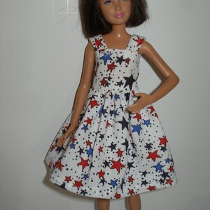 May include: A doll wearing a white dress with a pattern of red, blue, and black stars. The dress has a square neckline and a flared skirt. The doll has brown hair and is standing.