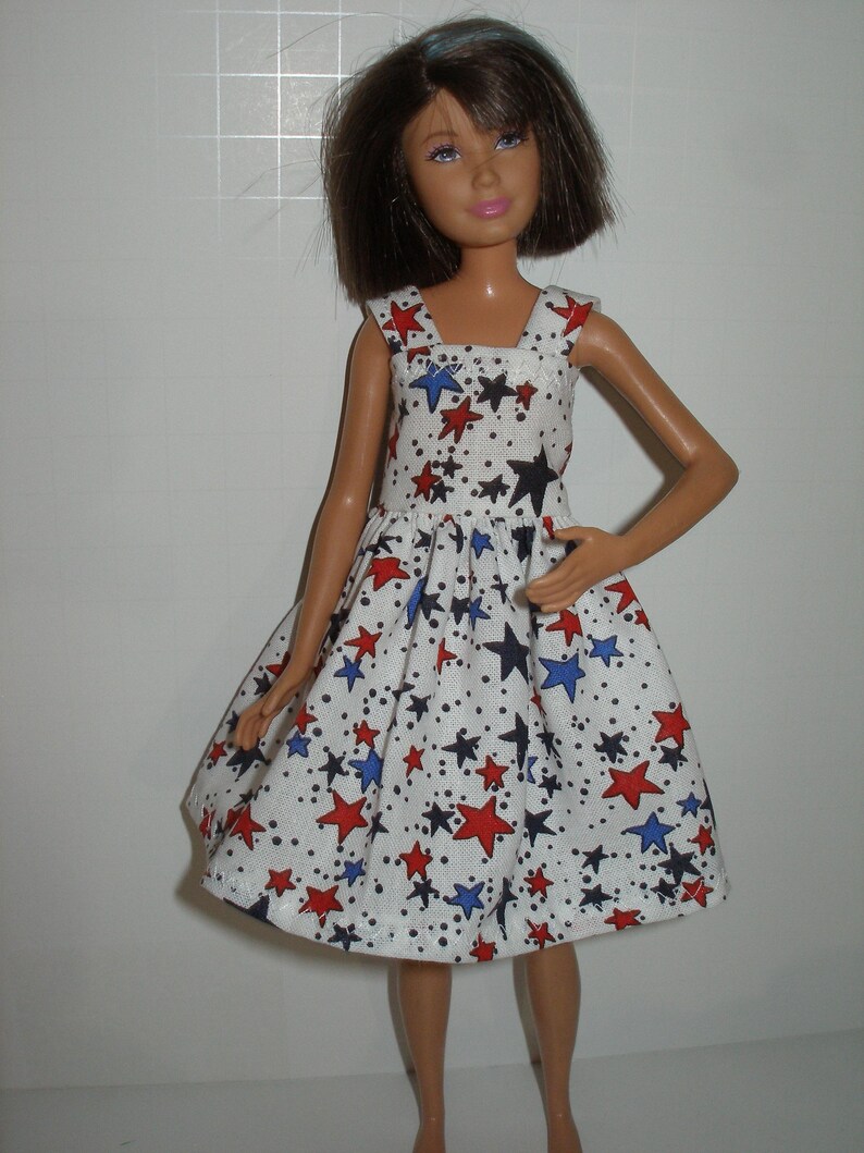 May include: A doll wearing a white dress with a pattern of red, blue, and black stars. The dress has a square neckline and a flared skirt. The doll has brown hair and is standing.