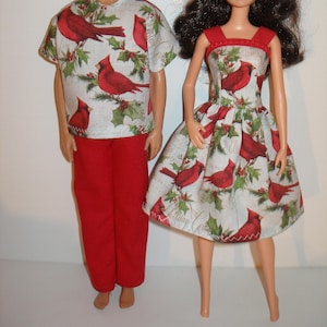 May include: Two dolls wearing matching outfits. One doll is wearing a red pants and a short-sleeved shirt with a pattern of red cardinals and green holly on a white background. The other doll is wearing a red-trimmed dress with the same pattern.