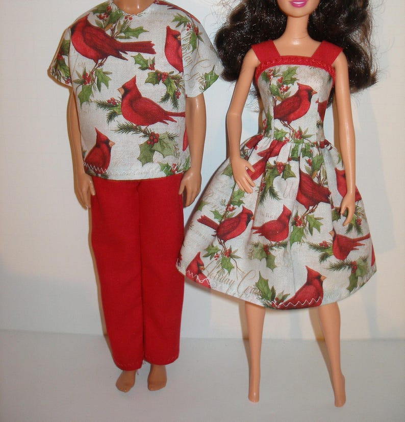 May include: Two dolls wearing matching outfits. One doll is wearing a red pants and a short-sleeved shirt with a pattern of red cardinals and green holly on a white background. The other doll is wearing a red-trimmed dress with the same pattern.