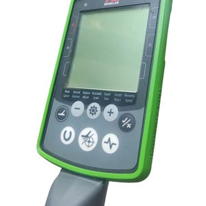 Puede incluir: Un detector de metales Minelab con un diseño verde y negro. El dispositivo tiene una pantalla y varios botones con iconos y texto. El mango es gris. El texto en el dispositivo incluye "Noise Cancel", "Volume Adjust" y "Target Tone".