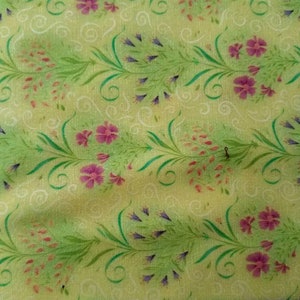 May include: A light green fabric featuring a repeating pattern of pink and purple flowers with green stems and white swirls. A purple measuring tape is positioned at the bottom, showing measurements from 9 to 13 inches.