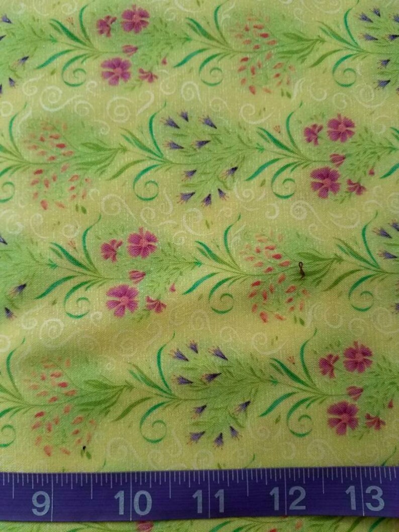 May include: A light green fabric featuring a repeating pattern of pink and purple flowers with green stems and white swirls. A purple measuring tape is positioned at the bottom, showing measurements from 9 to 13 inches.