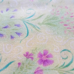May include: A light yellow fabric featuring a repeating pattern of purple and pink flowers, green leafy stems, and thin white swirling lines. The textile has a visible woven texture with a soft, matte finish across the entire surface area.