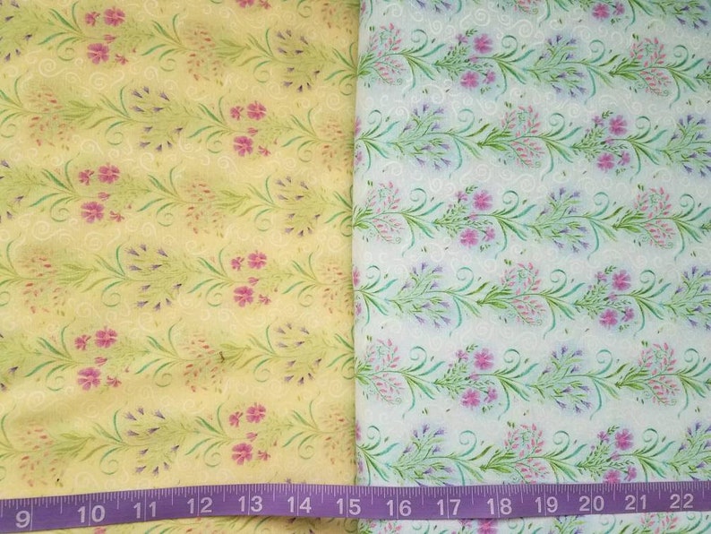 May include: Two pieces of fabric featuring a floral pattern with green vines and pink flowers. The left fabric has a yellow background, while the right fabric has a white background. A purple measuring tape is visible at the bottom, showing 22 inches.