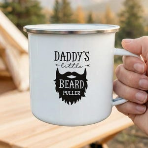 May include: White enamel camping mug with a silver rim, featuring the text "Daddy's little Beard Puller" in black. The mug has a black beard graphic. The mug is held in a hand, with a camping scene in the background.