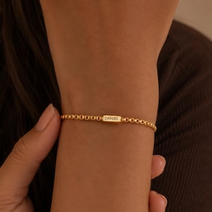 May include: A gold-toned box chain bracelet featuring a central rectangular bar charm engraved with the name DANIEL in capital letters. The bracelet has a uniform link design and a polished metallic finish, encircling the wrist with a simple clasp.