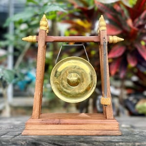 May include: A small, brass gong hangs from a wooden frame. The gong is circular with a central raised area. The frame is made of dark wood with decorative finials and a small mallet. The background is blurred with green and red foliage.