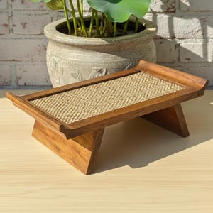 May include: A small, wooden serving tray with a woven, light brown surface. The tray has a rectangular shape with a raised border and angled legs. The wood has a warm, natural finish, and the tray is designed for serving or display.