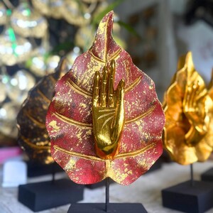 Pu&ograve; includere: Una scultura decorativa con una mano dorata che emerge da una foglia rossa con dettagli dorati. La foglia &egrave; montata su un supporto nero. Sono visibili altre sculture simili, suggerendo un tema di decorazione spirituale o artistica.