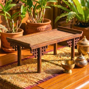 Può includere: Un piccolo tavolo rettangolare in legno con piano intrecciato e gambe decorative. Il tavolo è marrone e poggia su un runner fantasia. È circondato da piante in vaso e altri oggetti decorativi. Il tavolo è lungo circa 61 cm.
