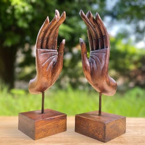 Op de afbeelding: Twee houten handsculpturen met open handpalmen, gemonteerd op metalen staven en vierkante houten bases. De handen zijn gesneden met gedetailleerde lijnen en rondingen, die de textuur van het hout laten zien. De sculpturen hebben een warme bruine kleur, tegen een vage groene achtergrond.
