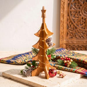 May include: A light brown wooden Christmas tree decoration featuring a star topper, wrapped in thin string lights. The tree is adorned with small pinecones, green holly leaves, and a single red glass ornament with gold painted details.