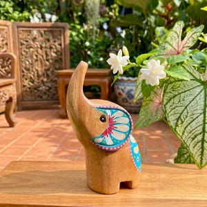 May include: Wooden elephant figurine with a raised trunk and painted floral designs in blue, red, and white. The elephant is standing on a wooden surface, with a natural background of plants and flowers.