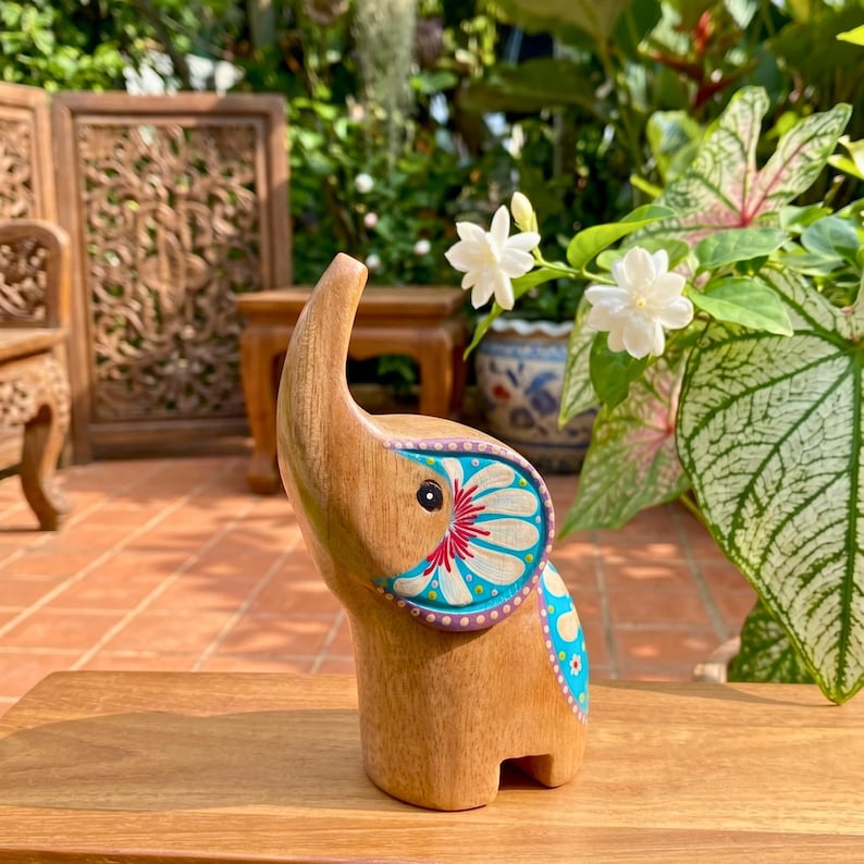 May include: Wooden elephant figurine with a raised trunk and painted floral designs in blue, red, and white. The elephant is standing on a wooden surface, with a natural background of plants and flowers.