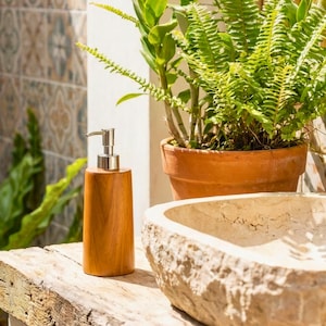 May include: A wooden soap dispenser with a silver pump sits on a weathered wooden surface. The dispenser is cylindrical and has a natural wood grain finish. A stone sink and potted plants are in the background, creating a spa-like atmosphere.