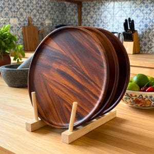 May include: Three round, dark brown wooden serving platters are displayed on a light-colored wooden rack. The platters have a rich wood grain pattern. The kitchen setting includes a mortar and pestle, cutting boards, and a bowl of limes.