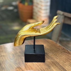 Può includere: Una scultura a forma di mano color oro, montata su una base quadrata nera con un'asta di metallo nero. La mano è in un gesto di offerta o ricezione. La scultura è probabilmente in metallo o resina ed è un oggetto decorativo.