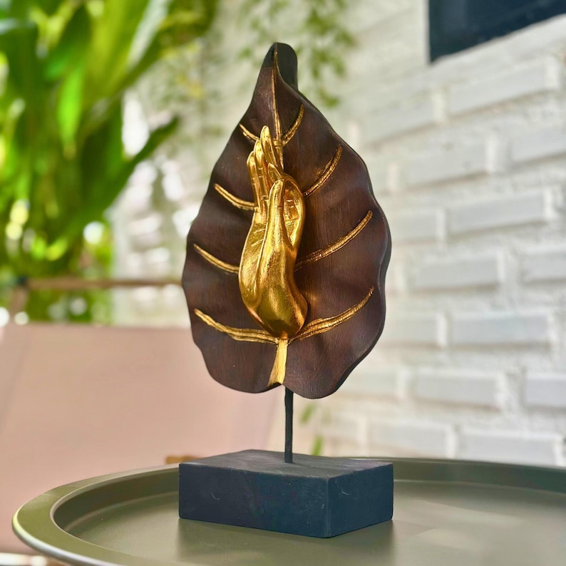 Pu&ograve; includere: Una scultura decorativa con una mano color oro che emerge da una foglia marrone scuro con dettagli dorati. La foglia &egrave; montata su un supporto in metallo nero e una base rettangolare nera. Un oggetto di arredamento spirituale o artistico.
