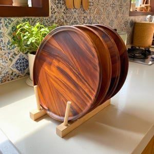 May include: A set of four round wooden plates with a rich, reddish-brown color, displayed in a light-colored wooden plate rack. The plates have a smooth surface and are stacked vertically. The rack has a simple, minimalist design.