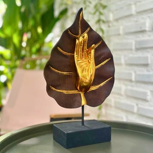 Pu&ograve; includere: Una scultura decorativa raffigurante una foglia marrone scuro con dettagli dorati e una mano dorata. La scultura &egrave; montata su una base e un supporto neri. Il design della foglia presenta venature dettagliate.