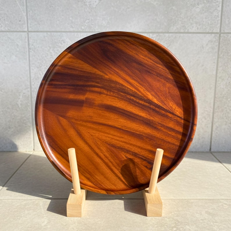 May include: A round, wooden serving tray with a rich, reddish-brown finish. The tray is displayed on a light-colored wooden stand. The wood grain is visible, adding texture to the surface. This tray is suitable for serving food or as a decorative piece.