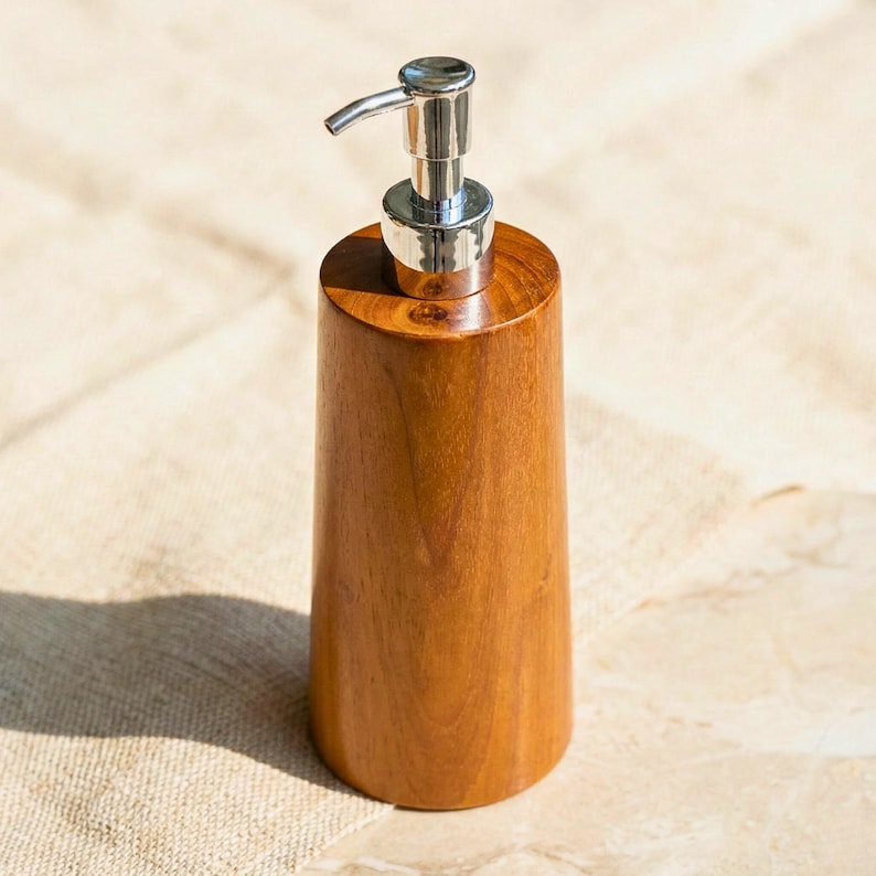 May include: A wooden soap dispenser with a silver pump. The dispenser is made of light brown wood and has a cylindrical shape. The pump is curved and has a shiny, metallic finish. The dispenser is sitting on a beige surface.