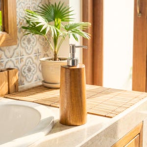 May include: A wooden soap dispenser with a silver pump top. The dispenser is cylindrical and made of light brown wood. It sits on a bamboo mat next to a white sink. A potted plant is in the background.