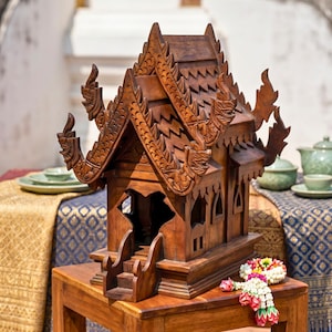 Può includere: Una casa degli spiriti in legno marrone scuro con intagli complessi, un tetto a più livelli con timpani appuntiti e decorazioni a forma di drago. La struttura presenta un ingresso aperto con piccoli gradini e una ghirlanda di fiori.
