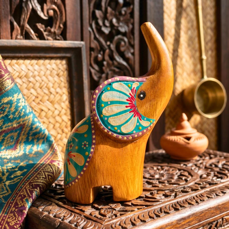 May include: A wooden elephant figurine with a trunk raised upwards. The elephant is painted with teal, white, and red floral designs on its ears and body. The figurine is displayed on a carved wooden surface.