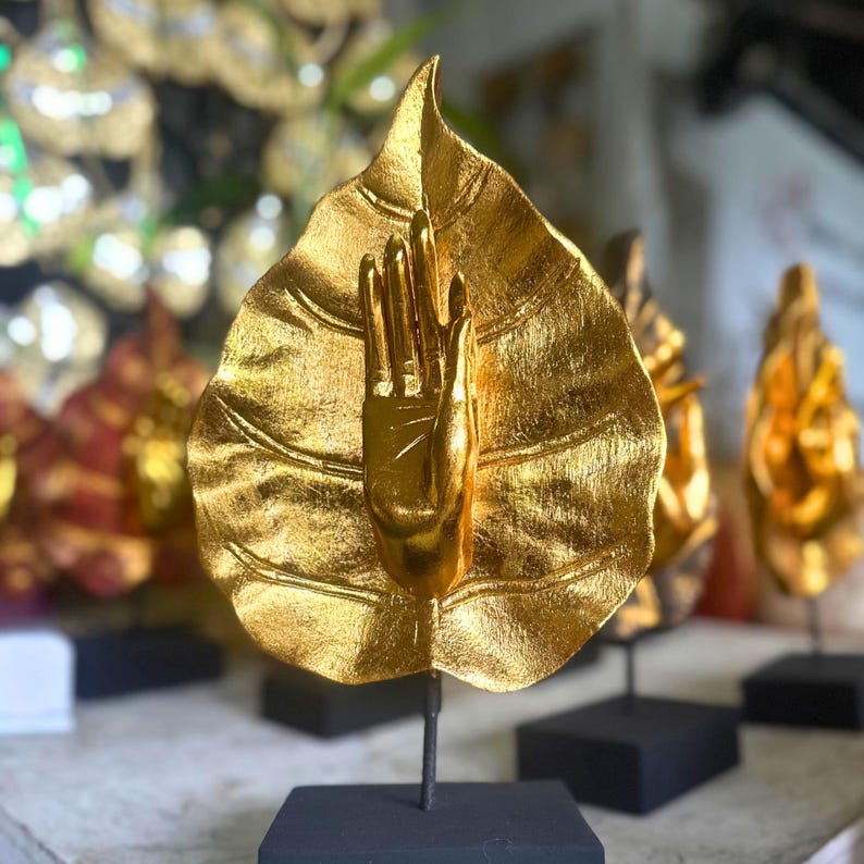 Pu&ograve; includere: Una scultura color oro di una mano che emerge da una foglia. La mano &egrave; alzata, con le dita estese. La foglia ha venature dettagliate. La scultura &egrave; montata su una base nera con un'asta di metallo. Altre sculture simili sono visibili sullo sfondo.