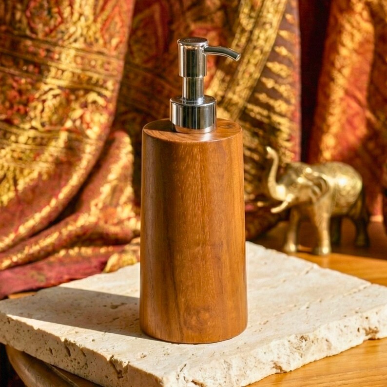 May include: A wooden soap dispenser with a silver pump. The dispenser is cylindrical and made of a warm-toned wood. The pump has a curved spout. A small gold elephant statue is in the background.