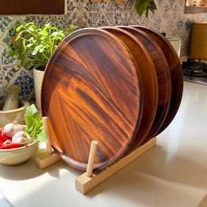 May include: A set of four round wooden serving platters with a rich, dark brown grain pattern. The platters are displayed in a light-colored wooden rack, showcasing their natural wood tones. The kitchen setting includes fresh herbs and spices.