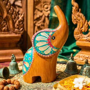 May include: Wooden elephant figurine with hand-painted turquoise, white, and red floral designs. The elephant has an upraised trunk and is set against a backdrop of carved wooden details and green fabric. Decorative bells and a wooden bracelet are in the foreground.