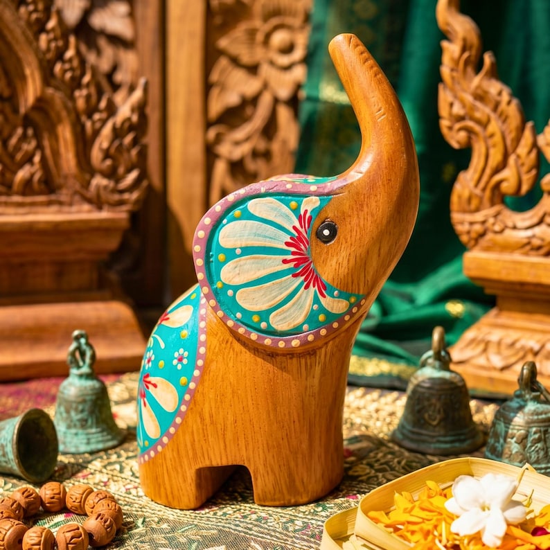 May include: Wooden elephant figurine with hand-painted turquoise, white, and red floral designs. The elephant has an upraised trunk and is set against a backdrop of carved wooden details and green fabric. Decorative bells and a wooden bracelet are in the foreground.