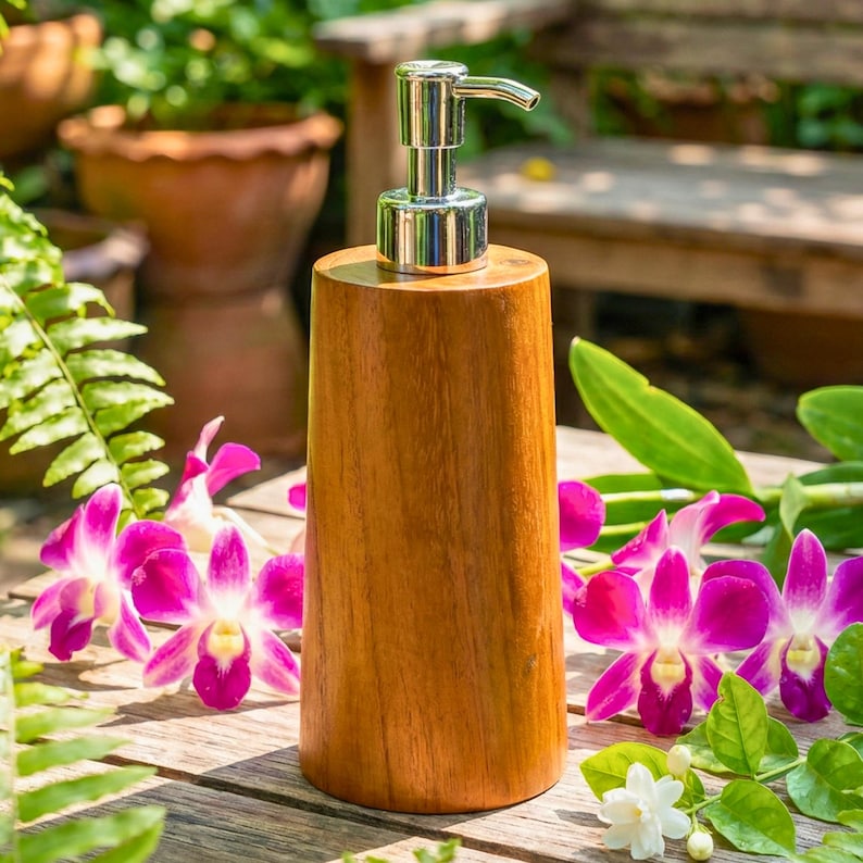 May include: A wooden soap dispenser with a silver pump. The dispenser is made of light brown wood and has a cylindrical shape. The background includes pink orchids and green foliage, creating a natural aesthetic.