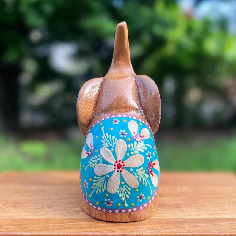 May include: Wooden elephant figurine with a brown body and a turquoise blue painted section. The blue section features a large white flower with red and yellow accents, surrounded by smaller flowers and green leaves. The elephant's ears are visible.
