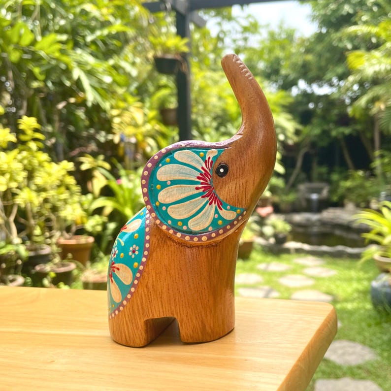 May include: A wooden elephant figurine with a trunk raised upwards. The elephant is painted with teal, white, and red floral designs on its ears and back. The figurine is brown and stands on a wooden surface.