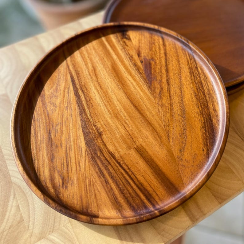 May include: Two round wooden serving trays with a rich, warm brown color and visible wood grain. The trays have a slightly raised edge and are likely made of acacia wood. One tray is in the foreground, and another is partially visible in the background.