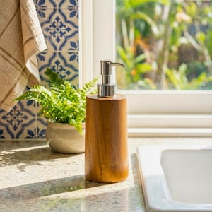 May include: A wooden soap dispenser with a silver pump. The dispenser is cylindrical and made of light brown wood. It sits on a speckled countertop near a white sink and a window with a view of greenery.