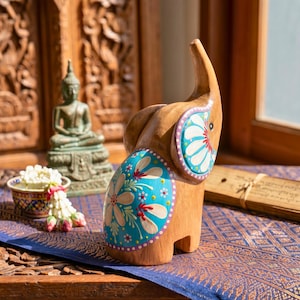 May include: Wooden elephant figurine with hand-painted turquoise and white floral designs. The elephant has a raised trunk and is set against a backdrop of carved wooden panels and a blue patterned textile. A small Buddha statue is visible in the background.