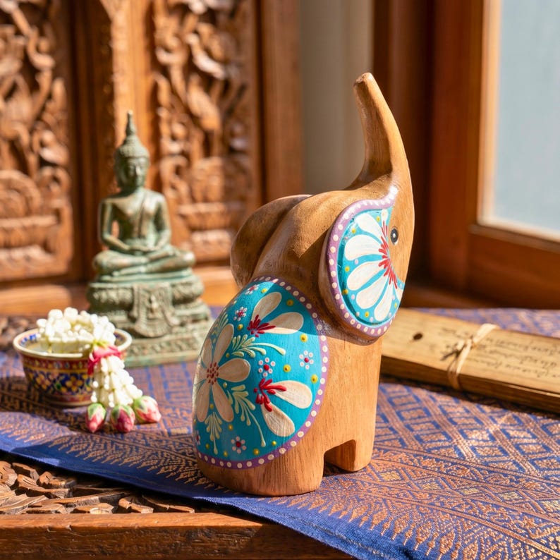 May include: Wooden elephant figurine with hand-painted turquoise and white floral designs. The elephant has a raised trunk and is set against a backdrop of carved wooden panels and a blue patterned textile. A small Buddha statue is visible in the background.
