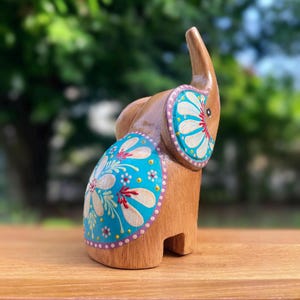 May include: Wooden elephant figurine with a trunk raised upwards. The elephant is painted with a light brown color and has blue and white floral designs on its sides and ears. The figurine is standing on a wooden surface.