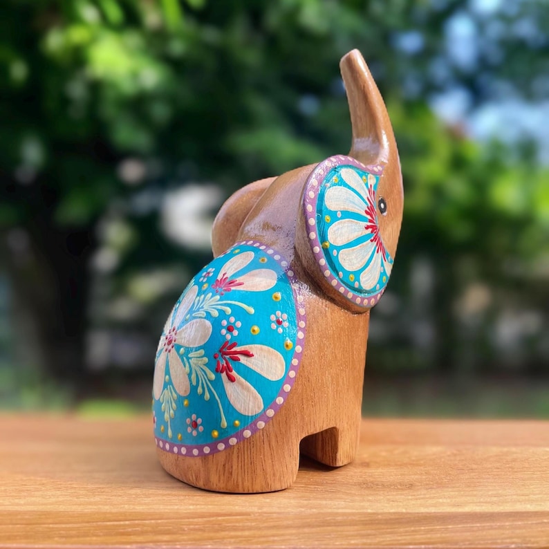 May include: Wooden elephant figurine with a trunk raised upwards. The elephant is painted with a light brown color and has blue and white floral designs on its sides and ears. The figurine is standing on a wooden surface.