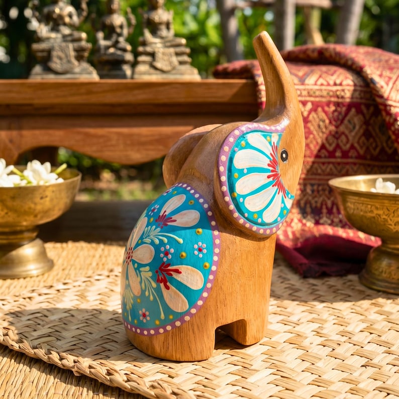 May include: Wooden elephant figurine with hand-painted turquoise and white floral designs. The elephant has a raised trunk and is decorated with purple and red accents. The figurine is displayed on a woven mat.