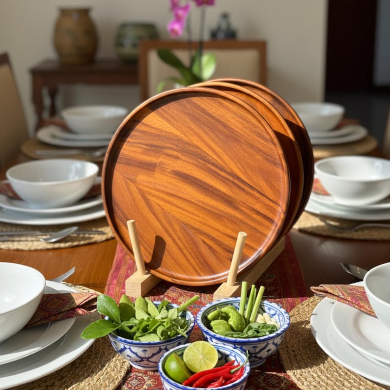 May include: A set of wooden serving trays with a rich, reddish-brown grain pattern, displayed on a wooden stand. The trays are part of a table setting that includes white bowls, plates, and small bowls filled with fresh ingredients like limes and chilies.