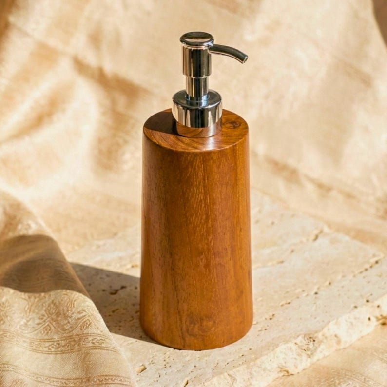 May include: A wooden soap dispenser with a cylindrical body and a silver pump. The dispenser is made of a light brown wood with visible grain. The pump has a curved spout. The dispenser sits on a light-colored stone surface.
