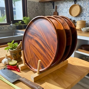 May include: A stack of four round wooden serving platters with a rich, reddish-brown grain pattern. The platters are displayed on a light-colored wooden stand. The kitchen setting includes a cutting board, knife, and fresh ingredients.