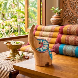 May include: A wooden elephant figurine with painted blue, red, and white details stands on a wooden table. The elephant has an upturned trunk and is next to a brass bowl and colorful pillows. The background includes a window and decorative items.